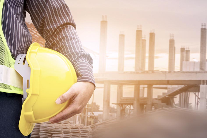 Brazo de persona que lleva un casco en la mano; detrás, planta en construcción