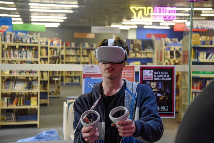 Un joven mira con lentes de realidad virtual; tiene objetos en sus manos; detrás hay una librería