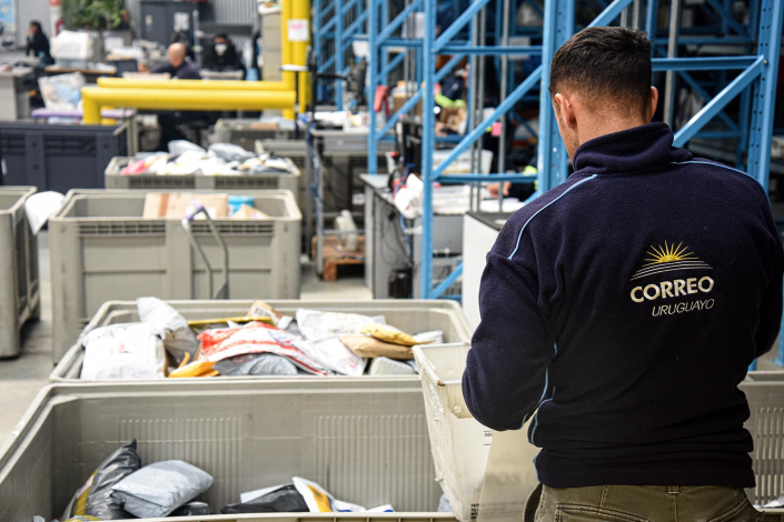 Persona con una campera del Correo Uruguayo, de espaldas, trabaja con cajas con objetos a su lado