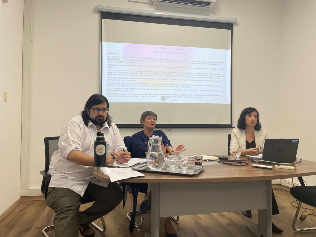 Tres personas sentadas detrás de una mesa y con una pantalla al fondo