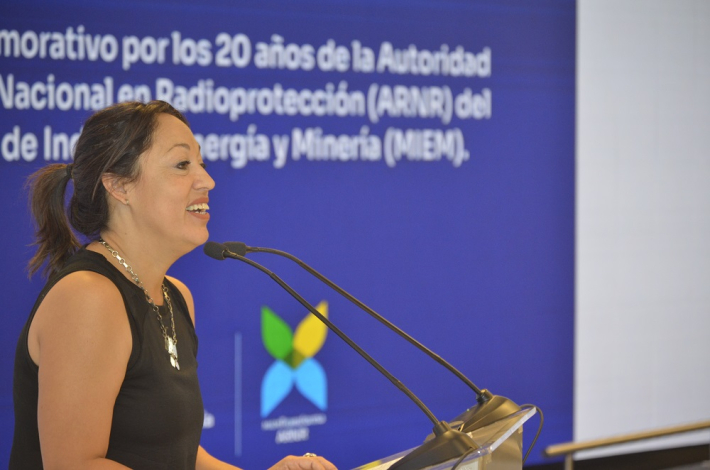Ministra Cardona de perfil habla a micrófono. Detrás, pantalla azul con logos y texto