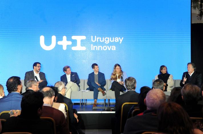 Público de espalda, sentado; al fondo, panel del evento; detrás, pantalla azul con logo del programa