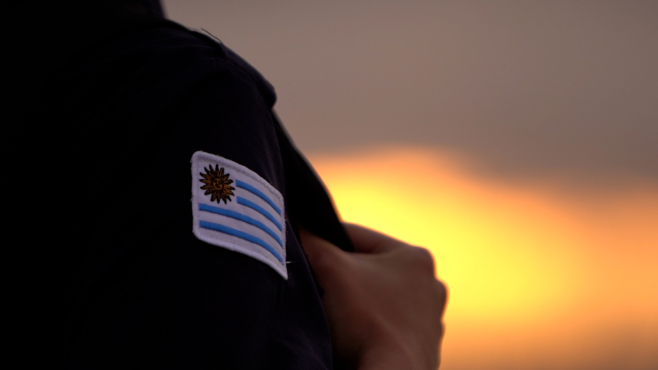 Primer plano de uniforme policial con bandera de Uruguay y atardecer de fondo con luz anaranjada.