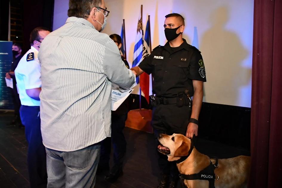 Acto de Reconocimiento a Policías destacados Un hombre le entrega un diploma a un policía, éste último está acompañado por un perro policía