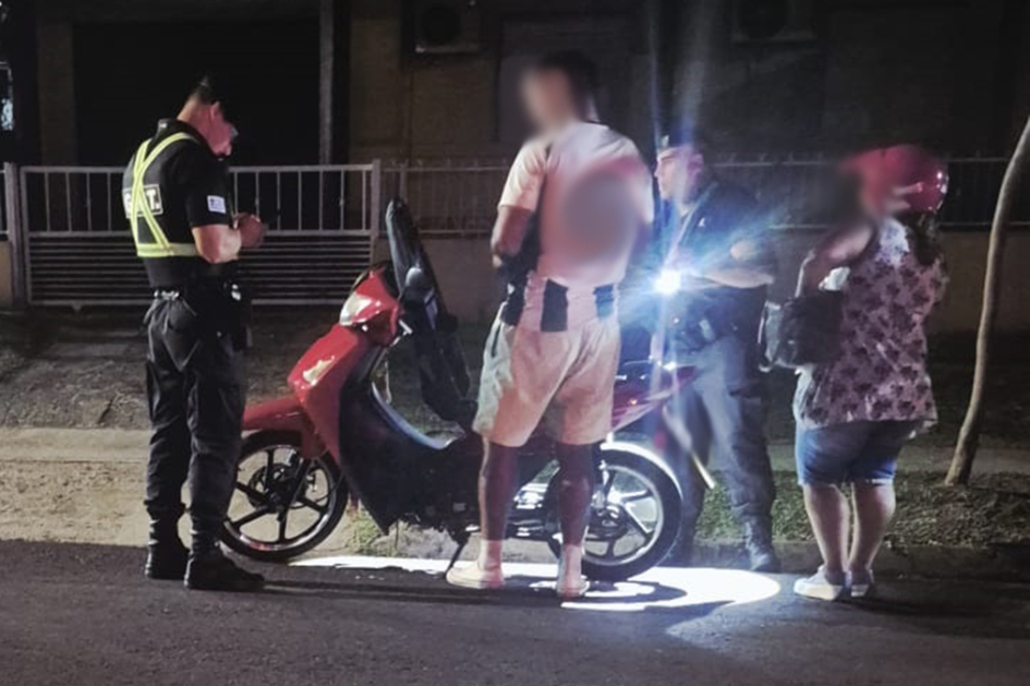 Dos personas en motocicleta siendo inspeccionados por dos policías.