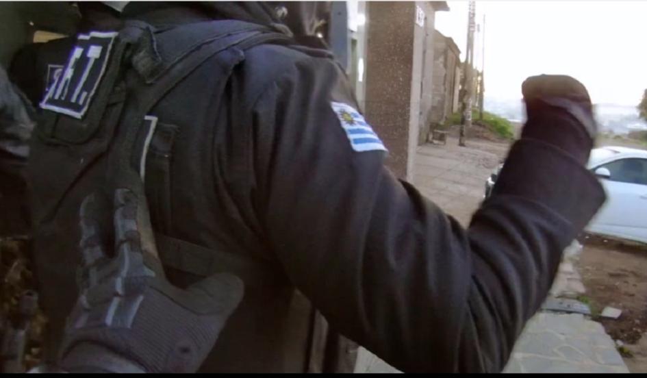 Policía de negro ingresando a allanamiento y su compañero lo impulsa con su mano izquieda de atrás Policías ingresando a allanamiento