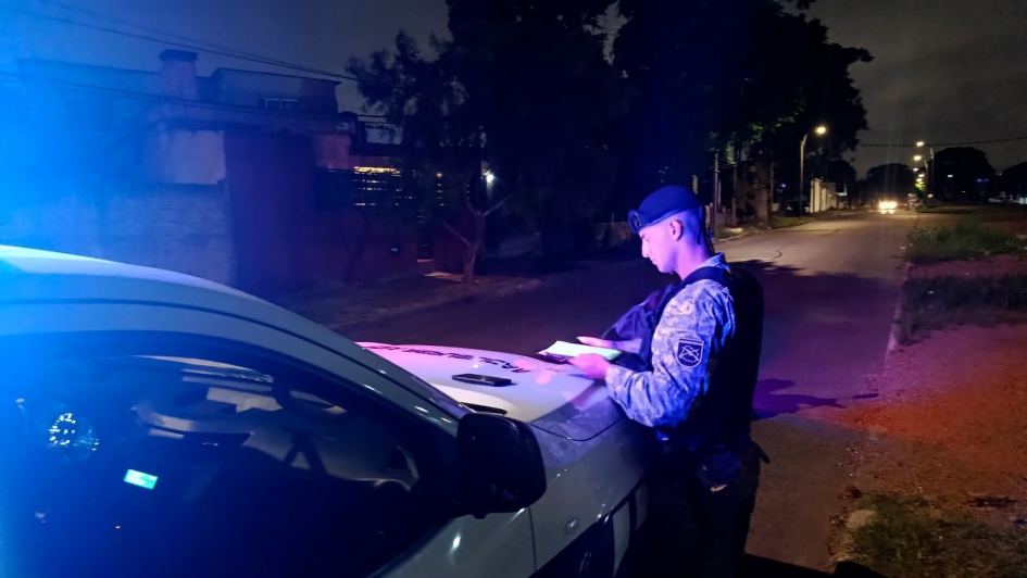 Policía toma notas apoyado en patrullero con luces encendidas en una calle nocturna.