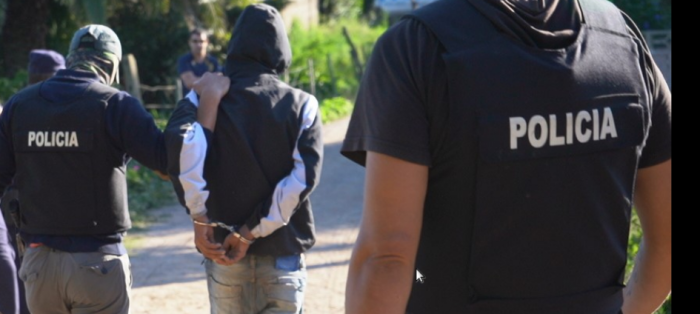 una persona con las manos esposadas en la espalda y dos policías al lado