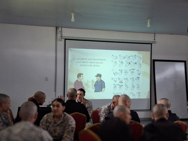 Policias sentados en sala de conferencias, de fondo una presentación con alfabeto dactilológico