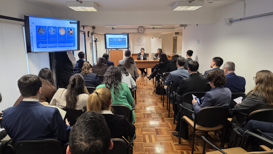 Personas sentadas en un salón viendo una presentación en pantalla. Personas sentadas frente a ellas,