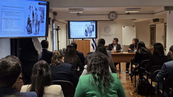 Personas sentadas en un salón viendo una presentación en pantalla. Personas sentadas frente a ellas,