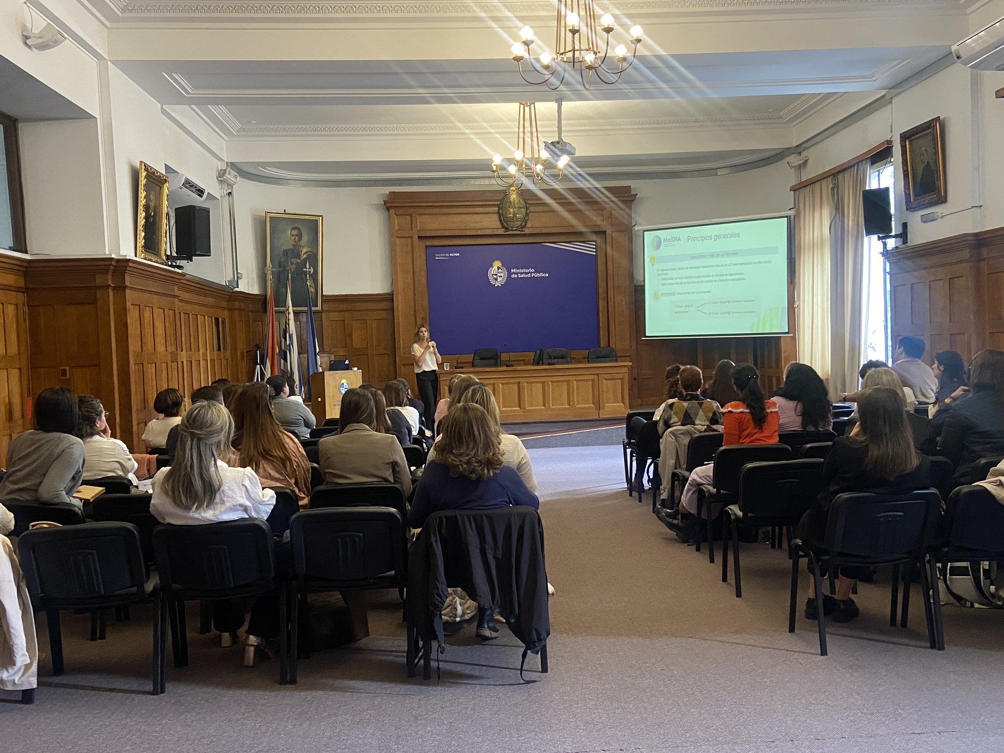 Participantes sentados en sillas, miran hacia persona que expone y pantalla.