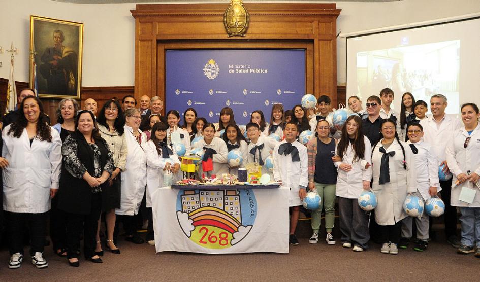 Foto grupal de autoridades con maestras y niños con túnica y moña. Al frente una maqueta de la playa