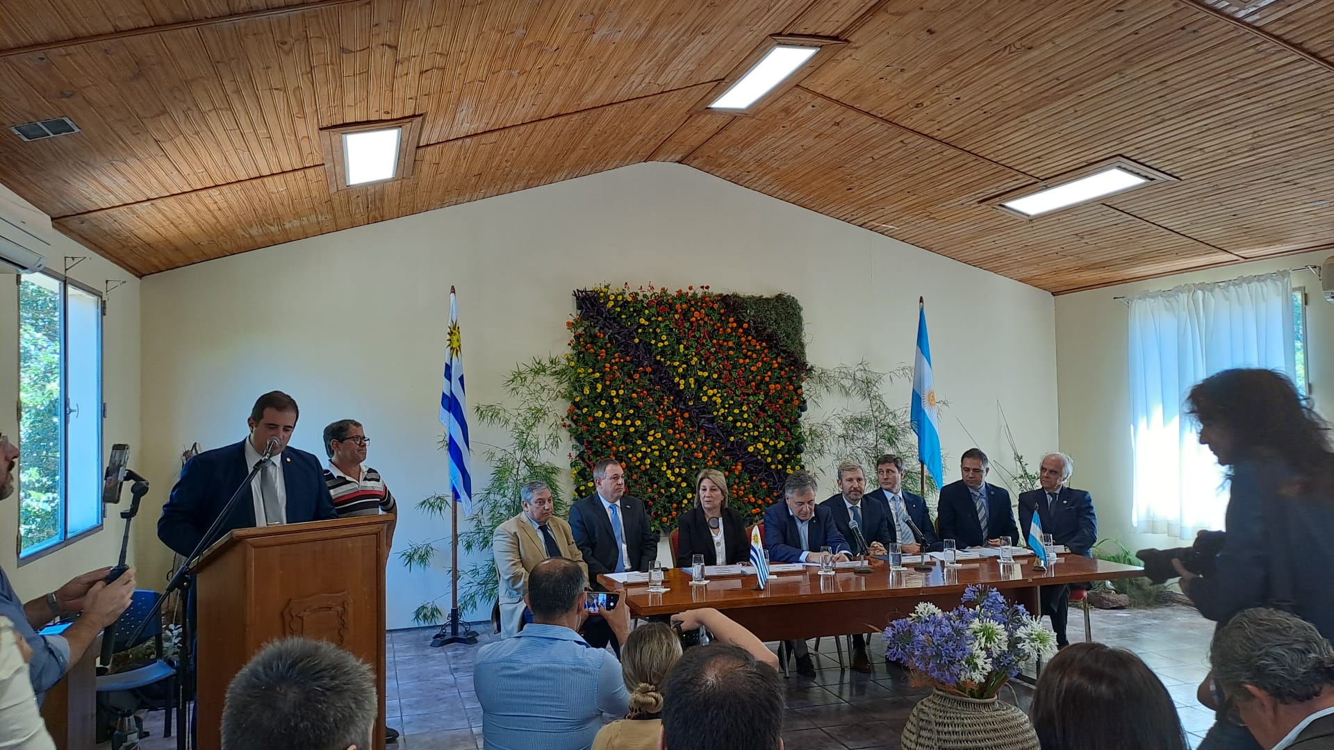 Mesa con 8 autoridades, una persona habla desde un atril. Al fondo banderas de Uruguay y Argentina