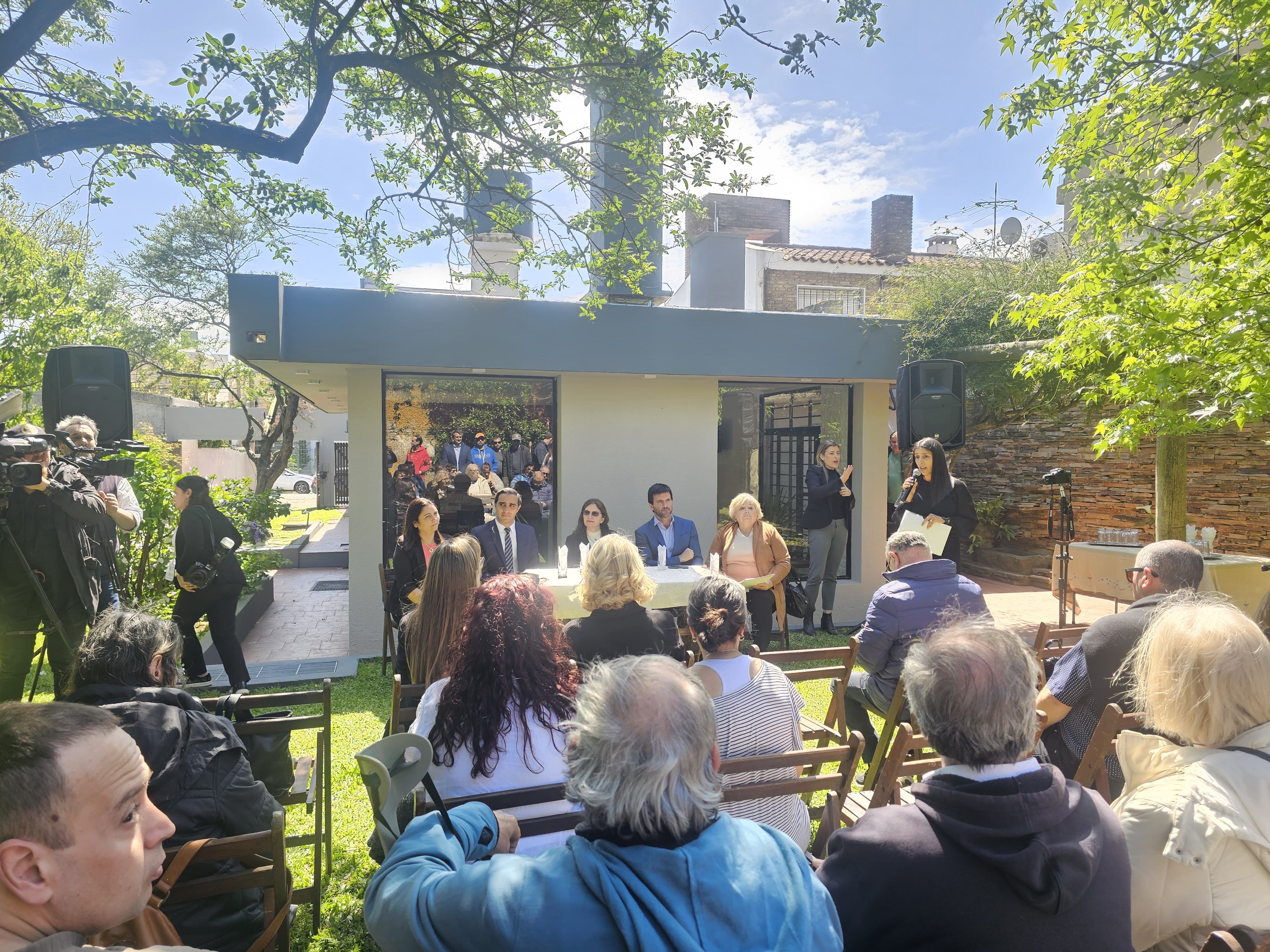 En el patio se ven los asistentes de espaldas, autoridades de frente, detrás, y al fondo la casa