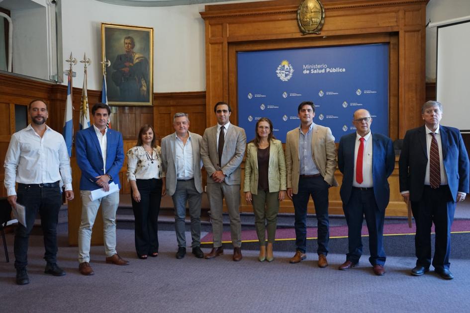 Firma de convenio para el tratamiento de tumores de cabeza y cuello Varias autoridades de pie, una al lado de la otra, en salón de actos, con fondo institucional