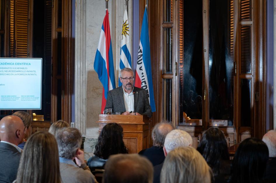 Briozzo expone en jornada académica Leonel Briozzo brinda discurso de pie. Detrás los pabellones patrios y al lado una pantalla