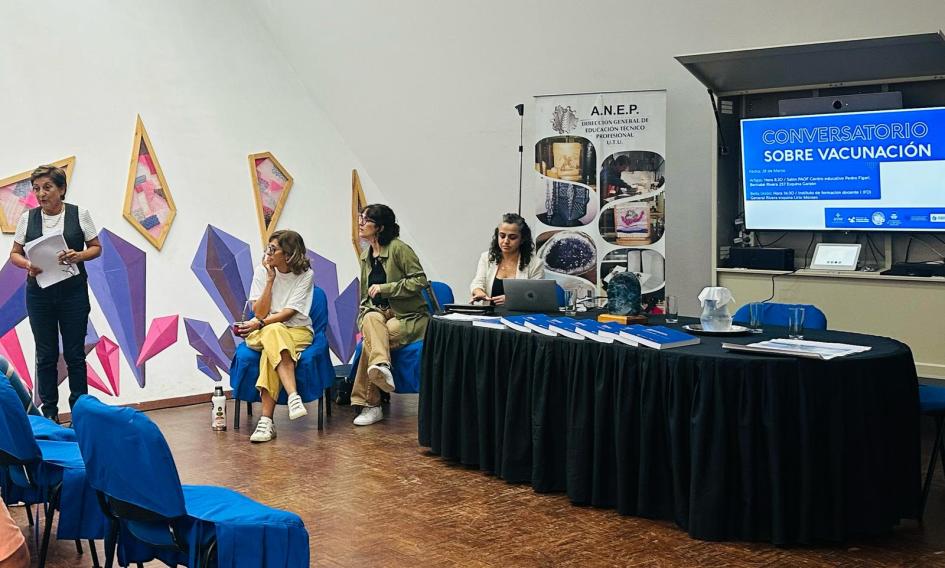 Expositoras del conversatorio sobre vacunación Una mujer expone de pie y tres sentadas la miran. Pantalla proyecta "conversatorio sobre vacunación"