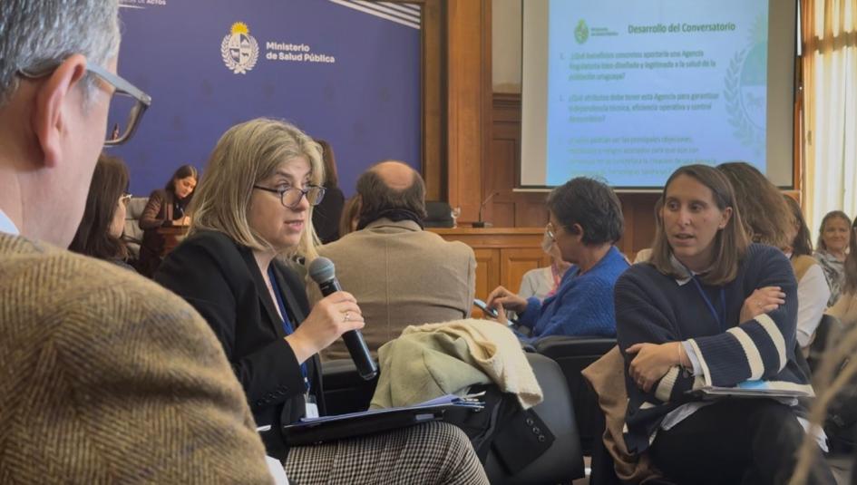 Puesta en común del trabajo grupal Una participante habla al micrófono, otros participantes de la ronda y auditorio la miran atentos