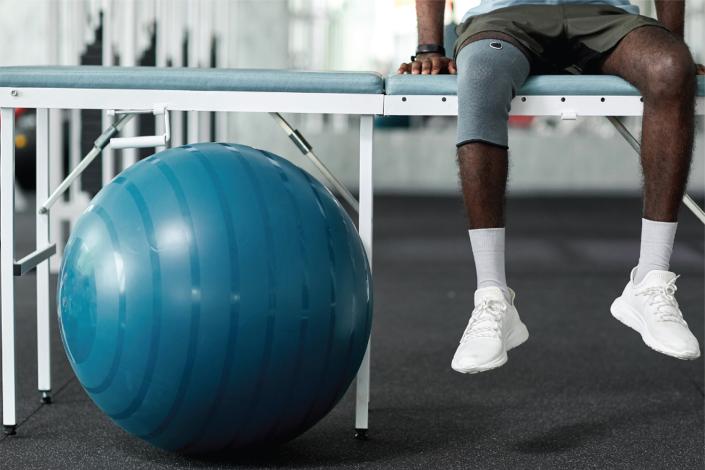 Piernas de un hombre sentado sobre una camilla. Tiene una rodillera. A su lado pelota de pilates.