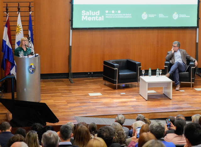 Ministra en escenario de pie detrás de un atril da un discurso al público sentado