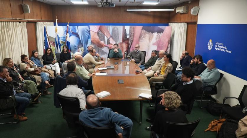 Mesa rectangular en el centro de una sala con personas sentadas al rededor, algunos en doble fila