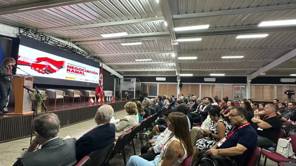 Imagen tomada desde un costado del salón donde se ven asistentes, atril con orador y la pantalla