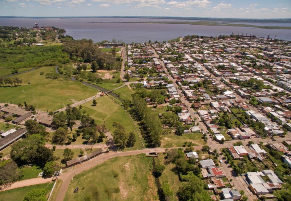 Fray Bentos, Río Negro Foto aérea de Fray Bentos donde se ve la ciudad con sus manzanas y vieviendas, el río Negro en el horizonte y el corte hacia la izquierda donde se encuentra una gran área verde que marca el fin de la ciudad.