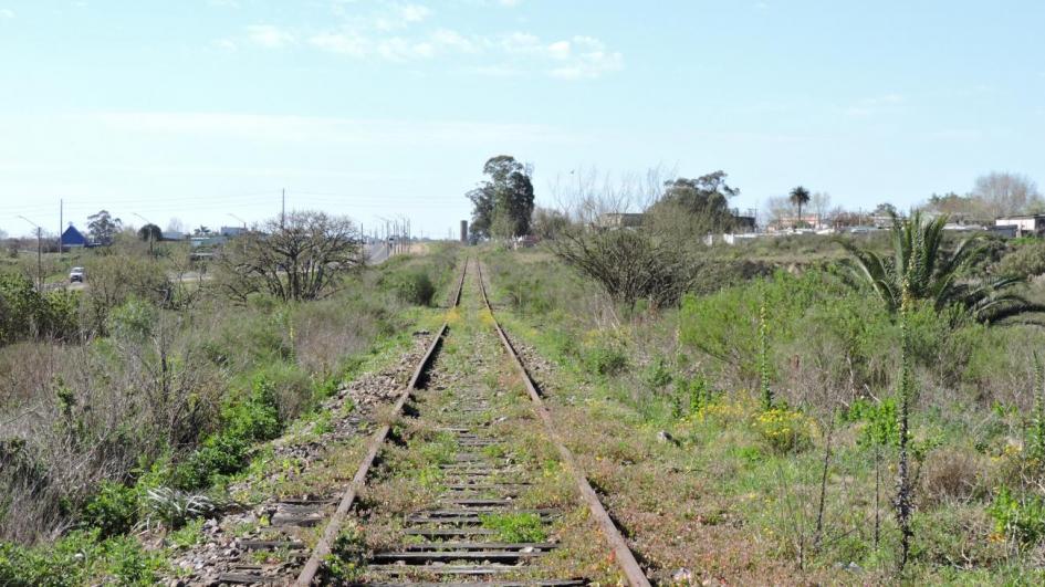Imagen que se ve en paralelo la ruta y las vías del tren, algunos vehículos por la ruta y en el horizonte al final del recorrido de las vías del tren edificaciones