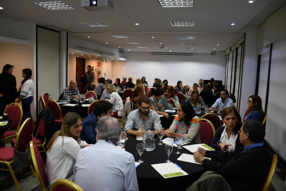 Panorama general de donde se realiza la actividad. Los y las asistentes a la actividad sentados en grupos en diferentes mesas