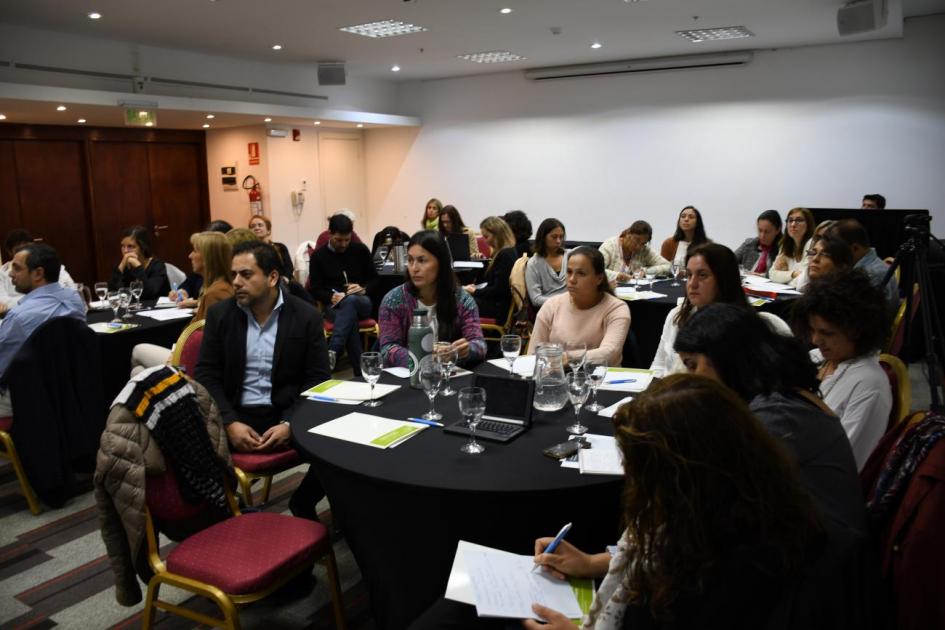 Panorama general de donde se realiza la actividad. Los y las asistentes a la actividad sentados en grupos en diferentes mesas