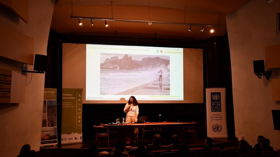 Foto de la especialista brasilera Emanoele Leal para en el escenario haciendo su presentación. Detrás se ve la pantalla gigante con una foto de la rambla de Curitiva con hombre haciendo ejercicio