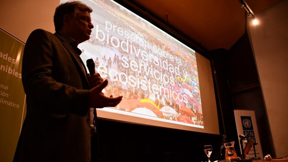 Otra foto de Fabio Scarano. En la pantalla de su presentación se proyecta una imagen de playa superpoblada con una leyenda por encima bien grande que dice: "Presión sobre la biodiversidad y servicios ecosistémicos"