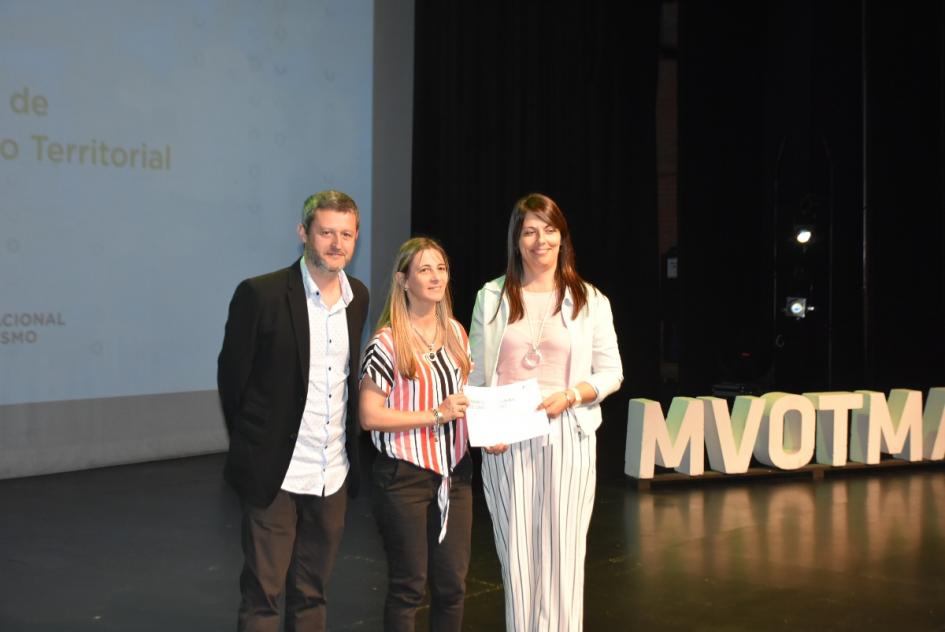 Miembro del jurado posa para la foto junto a dos de las ganadoras con el certificado en el escenario 