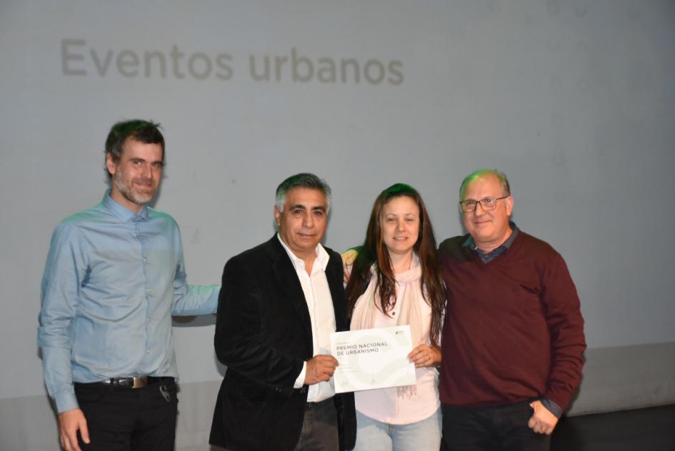 Miembro del jurado junto a una de las ganadores y dos de los ganadores posan para la foto en el escenario mostrando certificado 