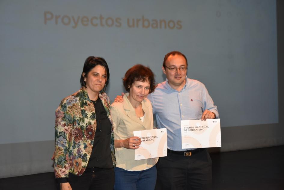 Una de las miembros del jurado posa para la foto junto a ganadora y ganador del Premio en el escenario y muestran sus certificados 