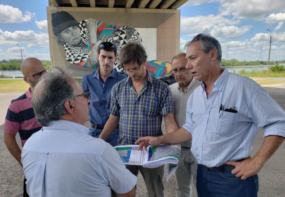 Recorrida rambla Mercedes El director nacional de Ordenamiento Territorial José Freitas está e la rambla de Mercedes en un día soleado junto a técnicos de la Dinot y la Intendencia parados en ronda. Freitas sostiene un mapa mientras que un técnico le explica. Atrás se ve un inmenso mural de una persona con ropa colorida