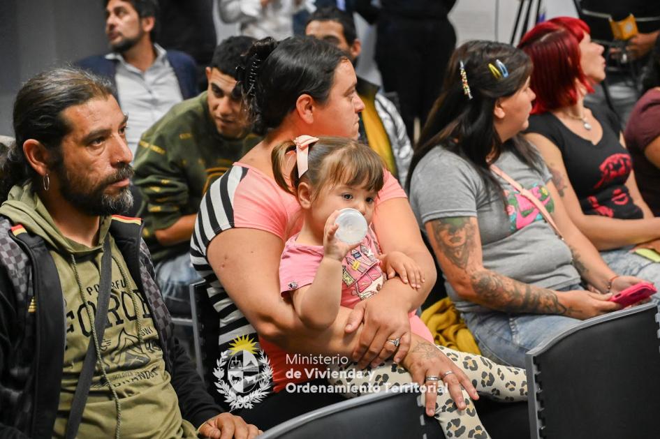 Gente en la firma del convenio Madre con hijo en brazos junto a otros beneficiarios sentados atendiendo a la firma del convenio