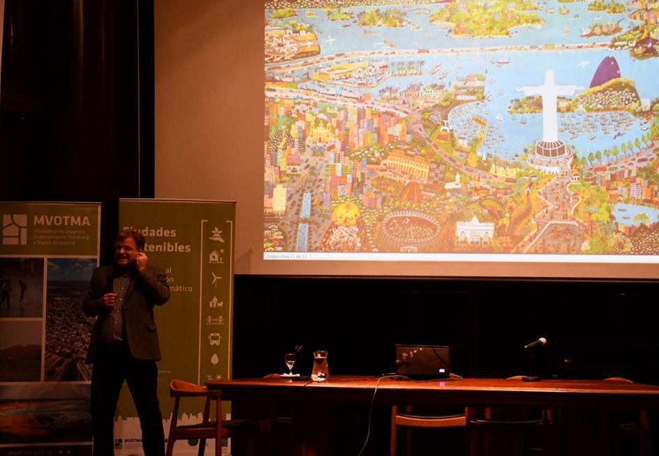 El técnico brasilero Fabio Scarano expone su presentación en el escenario. Detrás se ve en una pantalla gigante con su presentación que muestra una ilustración muy colorida de Río de Janeiro