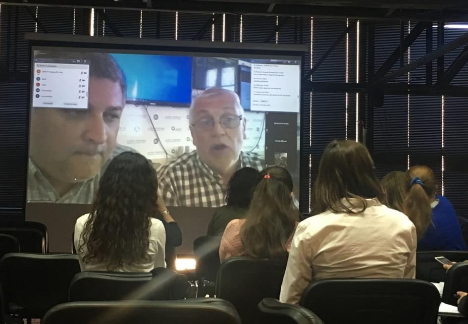 Reunión de trabajo donde se ve algunas técnicas de espaldas sentadas mirando a pantalla gigante donde se encuentran dos técnicos de otro país intercambiando a través de videollamada