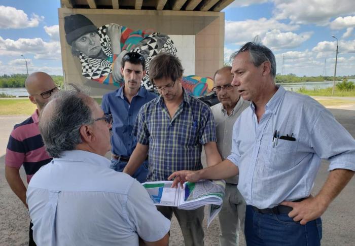 El director nacional de Ordenamiento Territorial José Freitas está e la rambla de Mercedes en un día soleado junto a técnicos de la Dinot y la Intendencia parados en ronda. Freitas sostiene un mapa mientras que un técnico le explica. Atrás se ve un inmenso mural de una persona con ropa colorida 