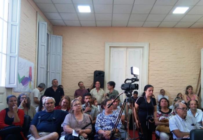 En una sala cerrada se encuentra variado público que escucha atento la presentación