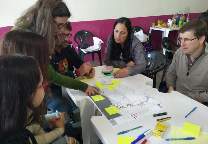 Taller con técnicos, vecinos y vecinas de la ciudad de Juan Lacaze. Sobre la mesa un mapa de la zona y papeles adhesivos. Alrededor de la mesa los participantes intercambiando.