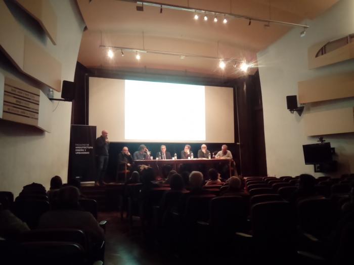  En el escenario de la sala de actos de FADU habla el decano Marcelo Danza parado y al costados sentados en el panel se encuentra el actual director nacional de Ordenamiento Territorial José Freitas, el director nacional de Vivienda Salvador Schelotto, los ex directores nacionales de Ordenamiento Territorial Roberto Villarmarzo y Manuel Chabalgoity