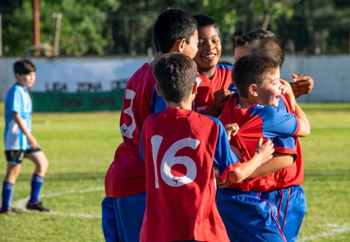 Cinco niños abrazados y sonrientes festejando un gol en cancha de fútbol. En segundo plano se ve otro niño de cuadro contrario caminando