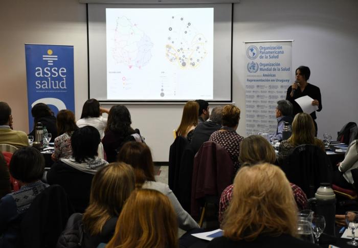 Se ve una técnica del Mvotma haciendo su presentación, pantalla gigante con varios mapas y personas sentadas en mesas circulares de espaldas mirando hacia la técnica que habla 