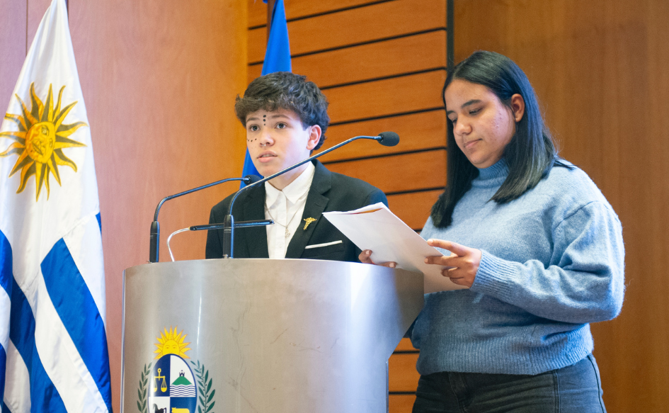 Dos adolescentes, integrantes del Consejo Asesor y Consultivo de INAU, parados detrás de un atril. Una de ellos lee un documento. Detrás, el pabellón nacional