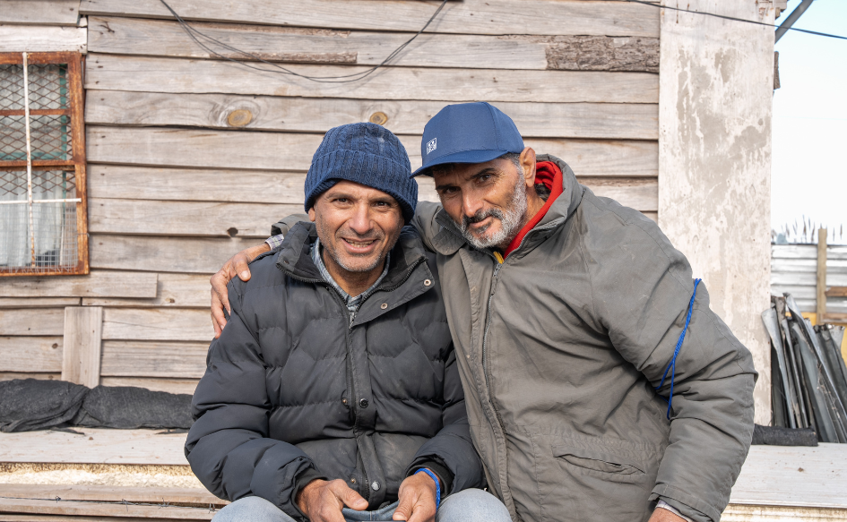 Dos vecinos de nuevo comienzo, abrigados con camperas y gorras, se abrazan y miran a la cámara. Detrás, una vivienda del barrio. 