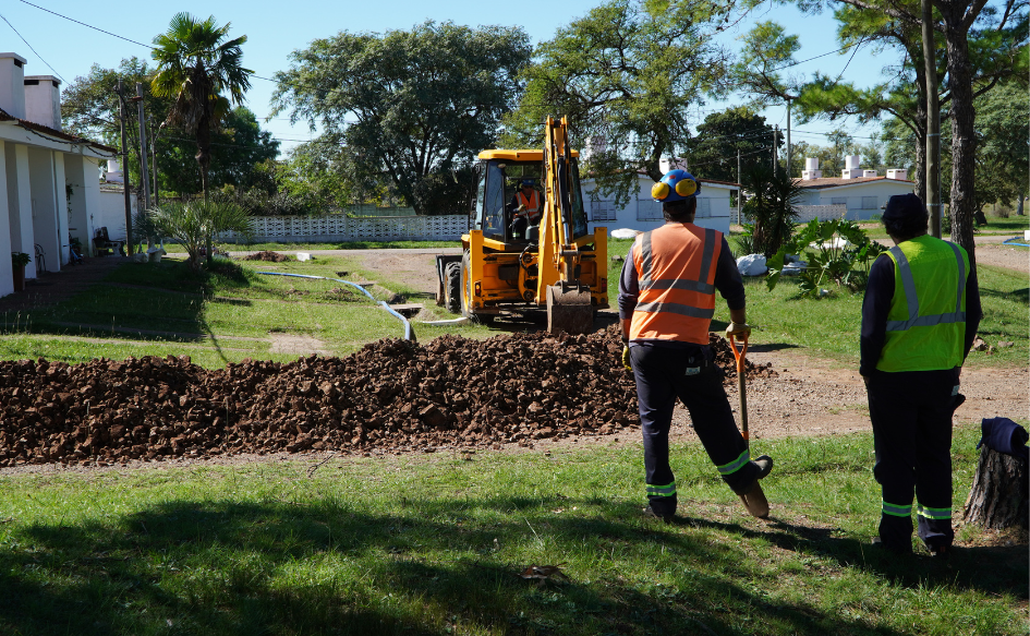 Retroexcavadora hace un pozo, mientras dos obreros, uno de ellos con una pala en la mano y el otro con las manos en los bolsillos, la observan