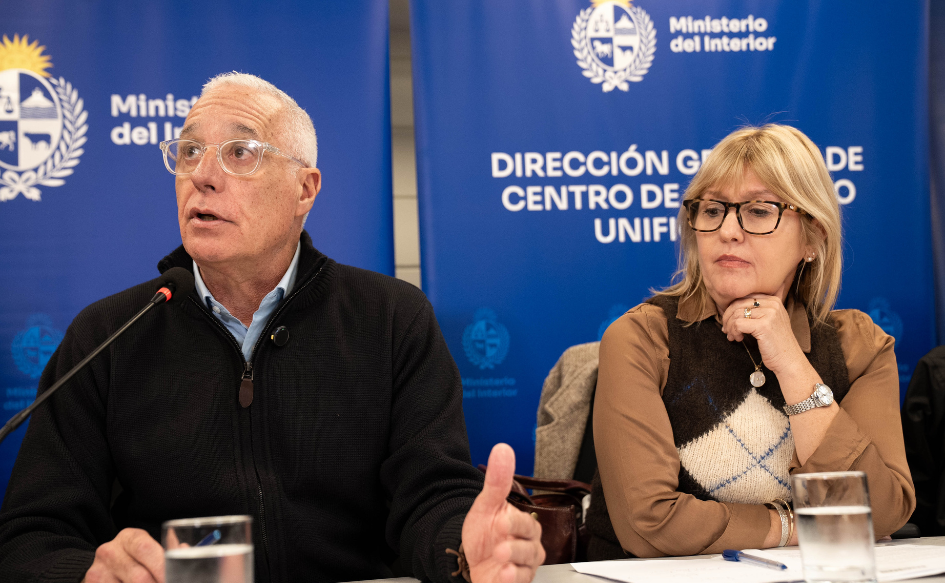 Ministro del Interior, Carlos Negro, habla en conferencia de prensa. A su lado, lo escucha atentamente la ministra de Salud Pública, Cristina Lustemberg. Detrás, banners con el logo del Ministerio del Interior
